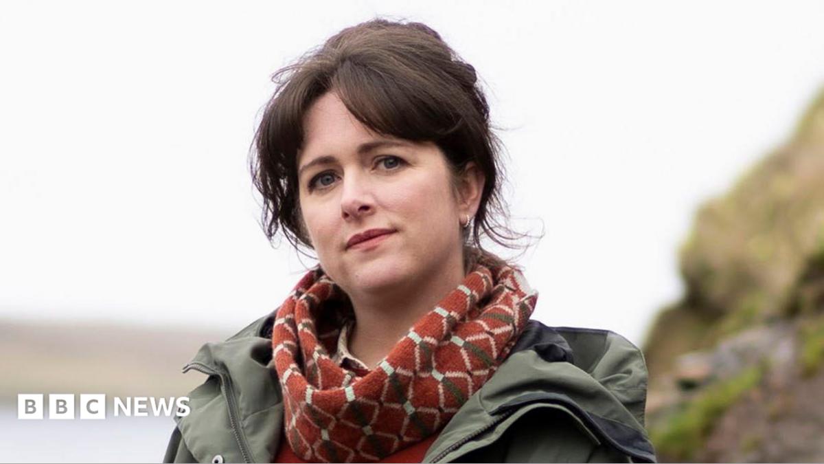 A young woman with dark hair stands on a rocky outcrop, next to open water. She is wearing a green jacket and a red scarf.