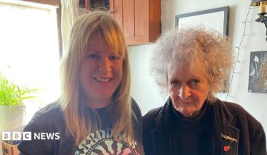A woman with long blonde hair and a fringe wearing a dark grey T-shirt stands next to an older woman who has short, curly grey hair and is wearing a brown jacket. They are both smiling at the camera.