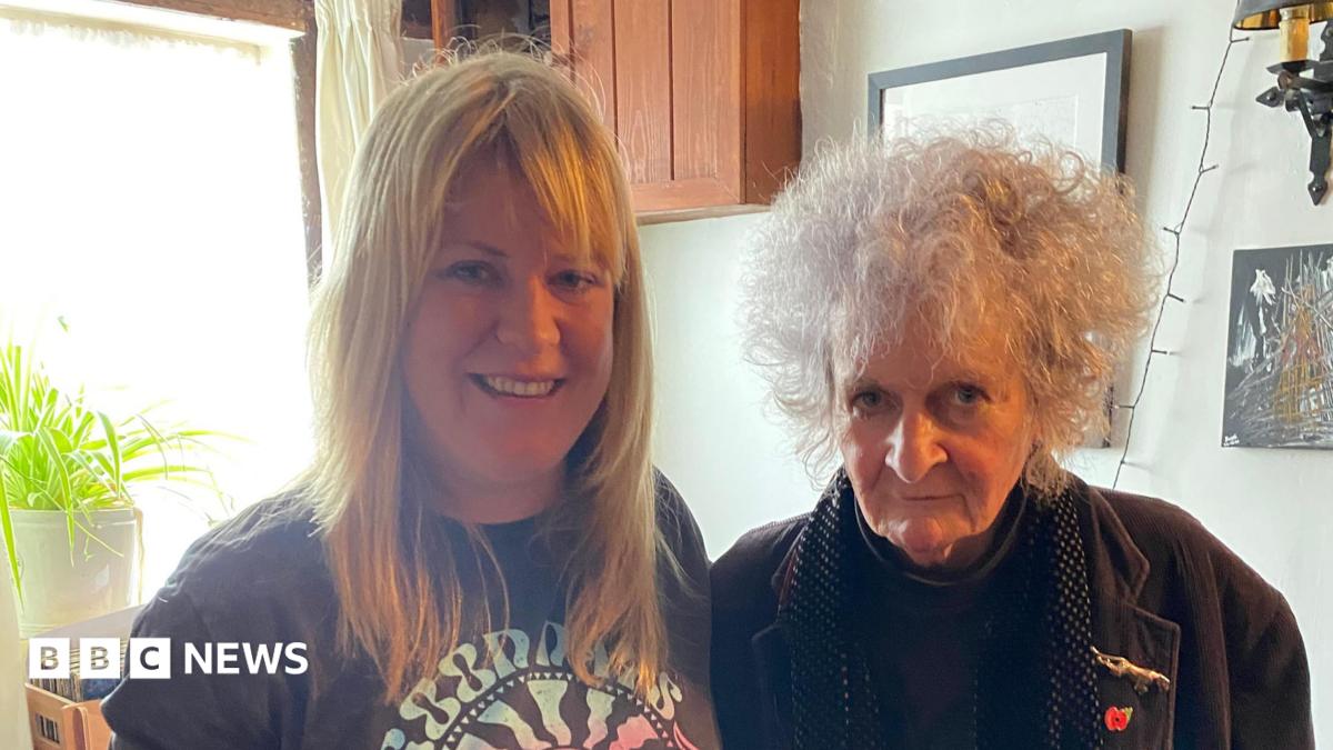 A woman with long blonde hair and a fringe wearing a dark grey T-shirt stands next to an older woman who has short, curly grey hair and is wearing a brown jacket. They are both smiling at the camera.
