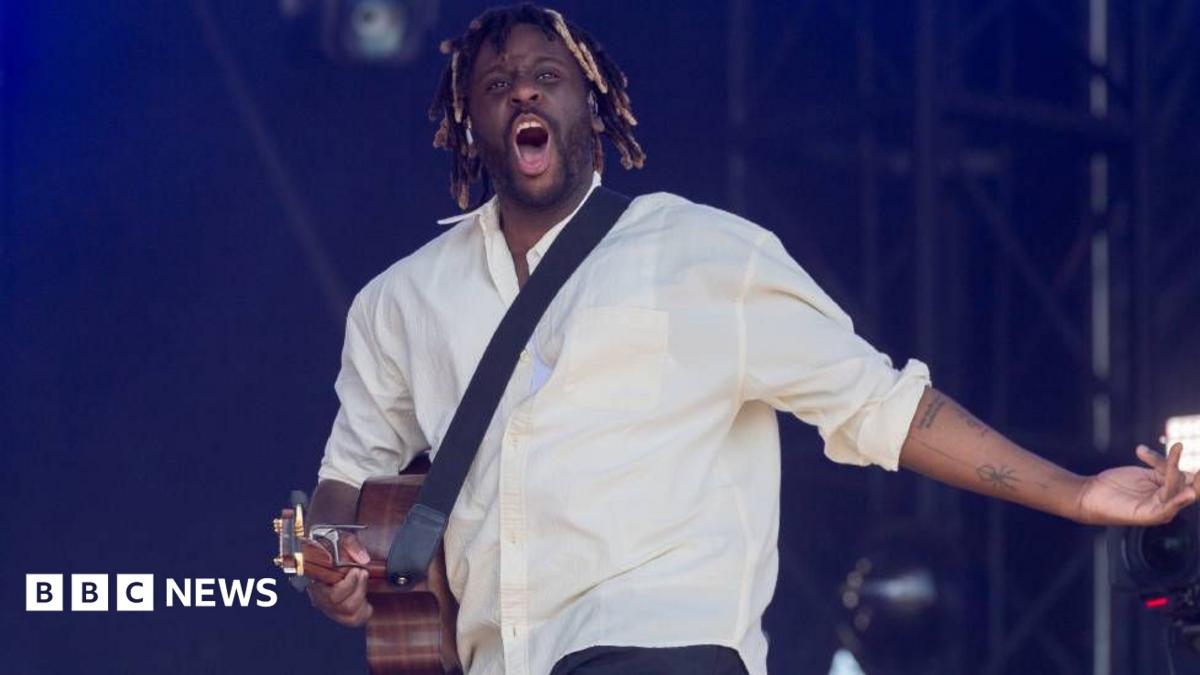 Myles Smith is wearing a white shirt and black jacket performing on stage at the 2026 MOBO Awards with a guitar and a microphone. The background behind him is orange.