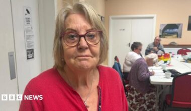 A woman with medium length hair is wearing red framed glasses and a red cardigan. She is standing in a community centre where three people are sat at a white table in the background.