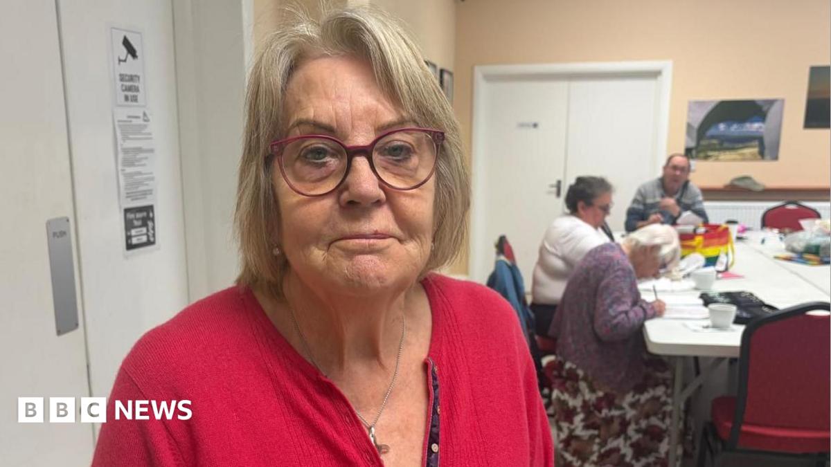 A woman with medium length hair is wearing red framed glasses and a red cardigan. She is standing in a community centre where three people are sat at a white table in the background.