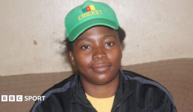 Madeleine Nseke Sissako, wearing a black tracksuit top over a yellow dress and a green baseball cap with the word cricket written on it in yellow, poses behind a table with a large collection of cricketing trophies, with some adorned with ribbons and one which says Africa Cup T20