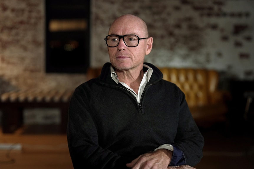 A man sitting indoors looks to his side, his hands on his lap. His expression is serious.