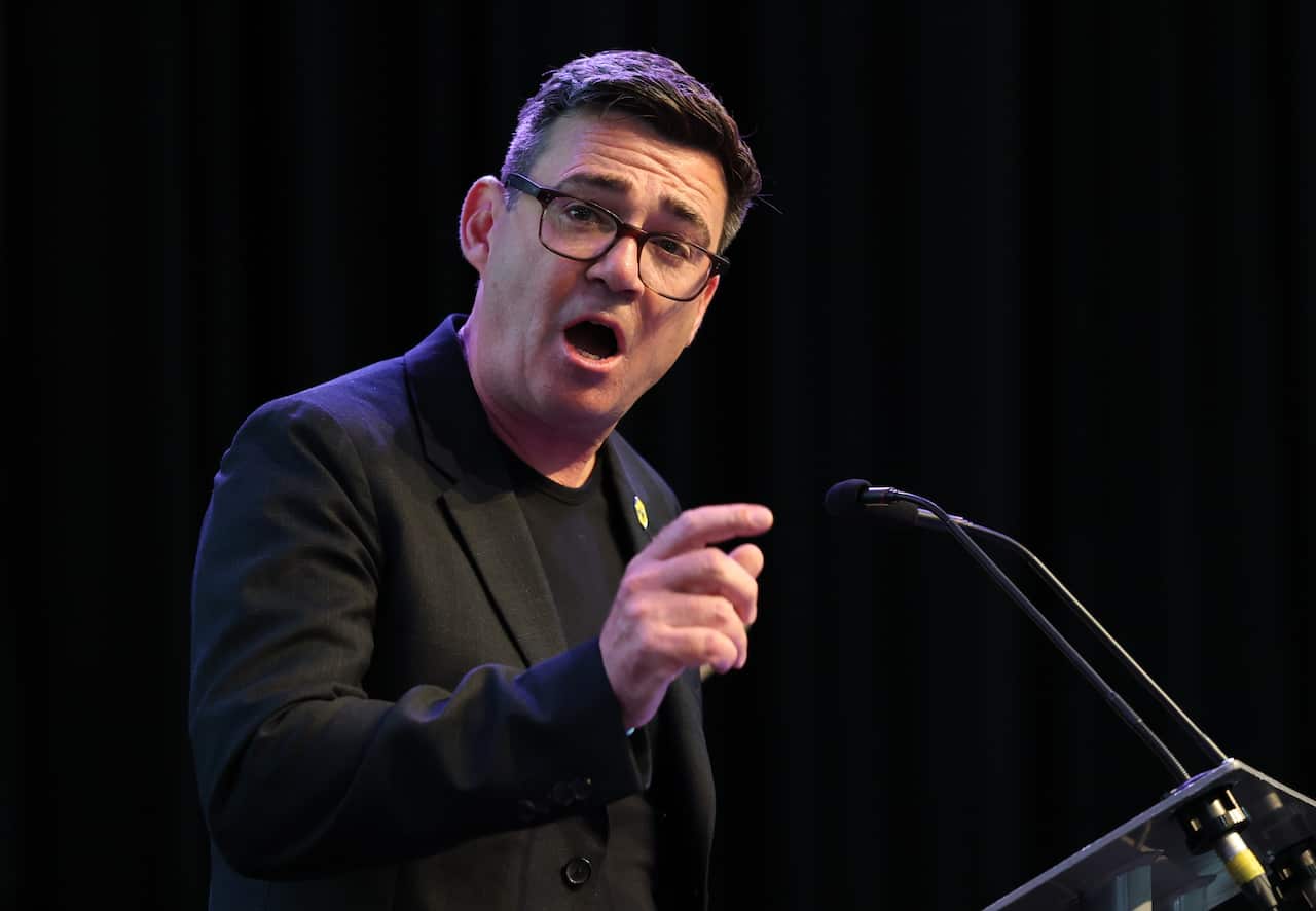 A bespectacled man in his mid-fifties dressed a black blazer and shirt speaks into a microphone.