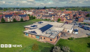 An aerial view of Thirsk and Sowerby Leisure and Wellbeing Hub, with solar panels installed on its roof. It's a large building with a mix of standard and flat roofing. There's a car park to the right of it, and a large expanse of grass around it with some trees. In the background there are residential streets, stretching off to trees and hills in the horizon. The sky is blue and cloudy.