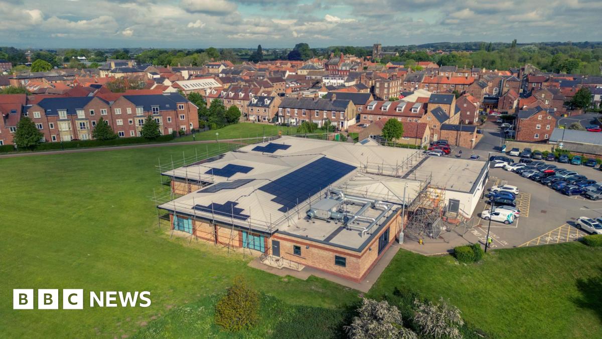 An aerial view of Thirsk and Sowerby Leisure and Wellbeing Hub, with solar panels installed on its roof. It's a large building with a mix of standard and flat roofing. There's a car park to the right of it, and a large expanse of grass around it with some trees. In the background there are residential streets, stretching off to trees and hills in the horizon. The sky is blue and cloudy.