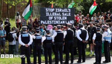Eight police officers watching on as people protest on a field of grass. There are four Palestinian flags being waved and a large sign being held up with the words 'STOP ISRAEL'S ILLEGAL SALE OF PALESTINIAN LAND'.
