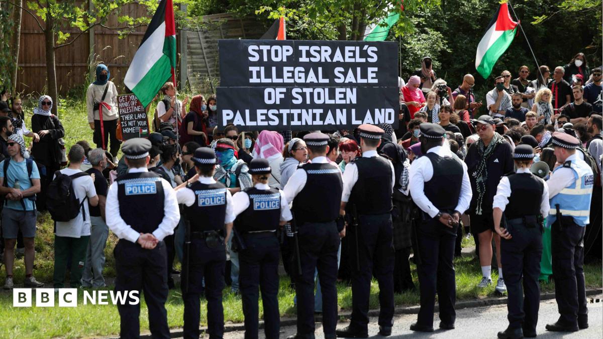 Eight police officers watching on as people protest on a field of grass. There are four Palestinian flags being waved and a large sign being held up with the words 'STOP ISRAEL'S ILLEGAL SALE OF PALESTINIAN LAND'.
