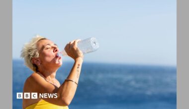 A woman with short blonde hair drinks from a plastic water bottle by the sea.