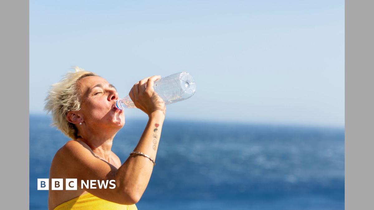 A woman with short blonde hair drinks from a plastic water bottle by the sea.