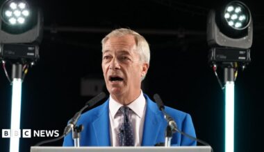 Nigel Farage in a bright blue suit, white shirt and black tie, with red and white coloured dots stands behind a lectern. Two lights are either side of him.
