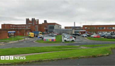 Furness General Hospital in Barrow. Buildings with orange bricks are interspersed with roads and grassy banks. Ambulances are parked to one side.