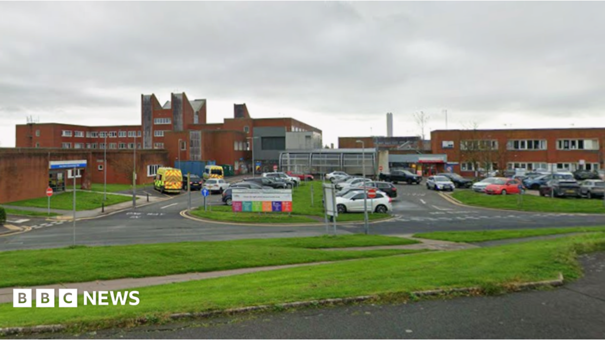 Furness General Hospital in Barrow. Buildings with orange bricks are interspersed with roads and grassy banks. Ambulances are parked to one side.