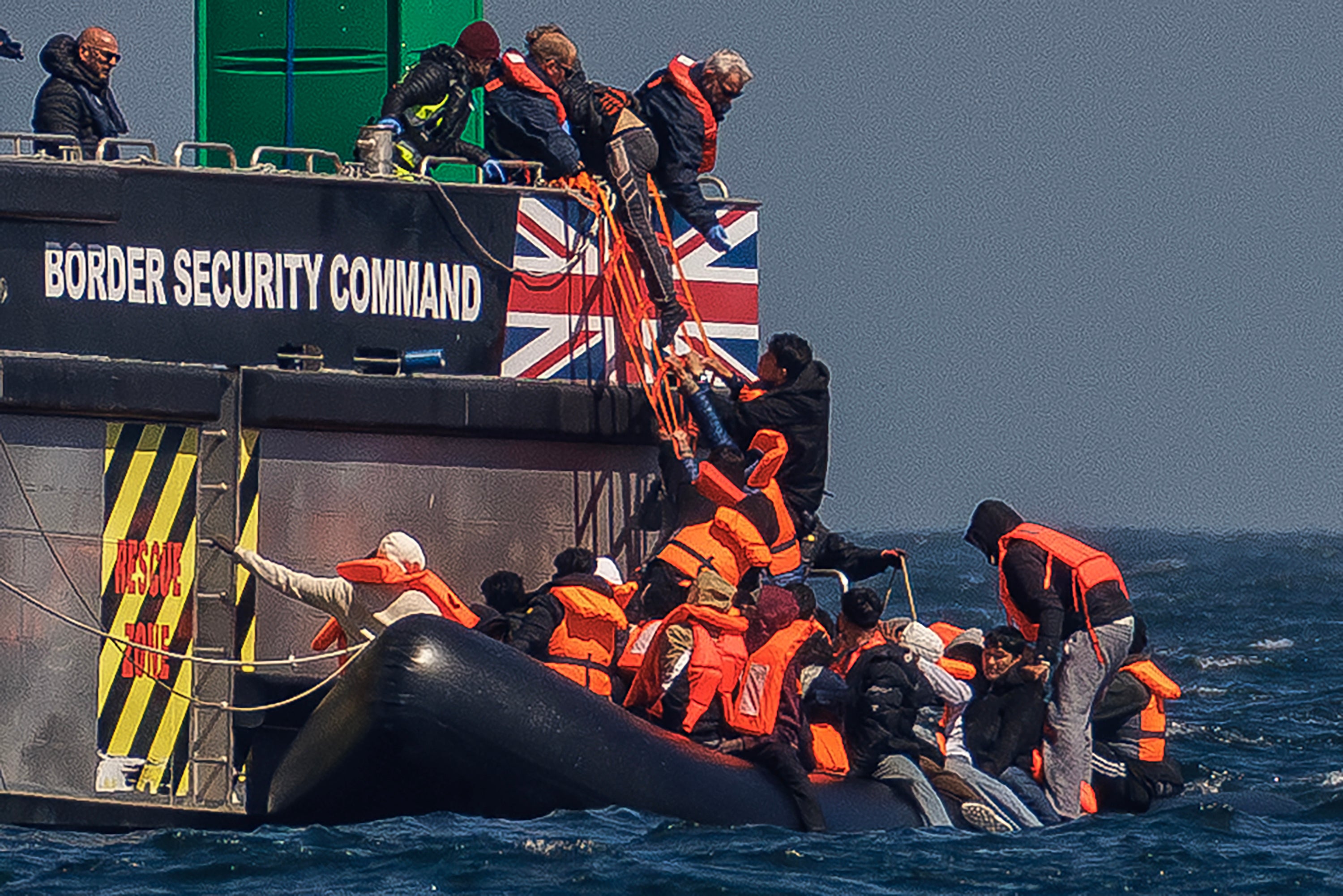 Migrants on an inflatable 'small boat' clamber on to The UK Border Force vessel, BSC Intrepid after crossing the channel from northern France on April 27, 2026 in Dover, England. AI facial age estimation technology will be used on migrants at the UK border.