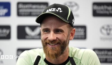 Kane Williamson smiling and wearing a black New Zealand cap sits in front of two long black microphones