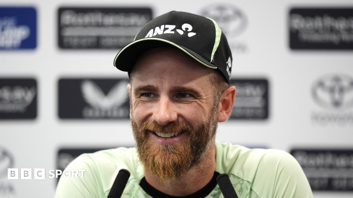 Kane Williamson smiling and wearing a black New Zealand cap sits in front of two long black microphones
