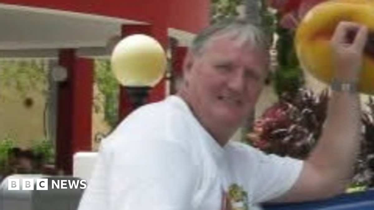 A slightly blurry picture of a man in his 60s with grey hair, wearing a white t shirt.