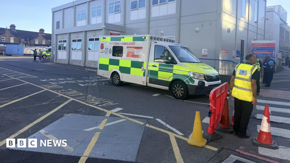 An ambulance outside a hospital block, that is beige in colour. People with high-vis jackets are to the right, and traffic cones and a red plastic barrier, by a crossing. Other people are standing to the left, by another emergency vehicle.
