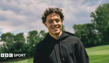 A photo of Isaac Heath smiling at the camera with trees in the background. He is wearing a black hoodie with the Cambridge United club badge on the chest