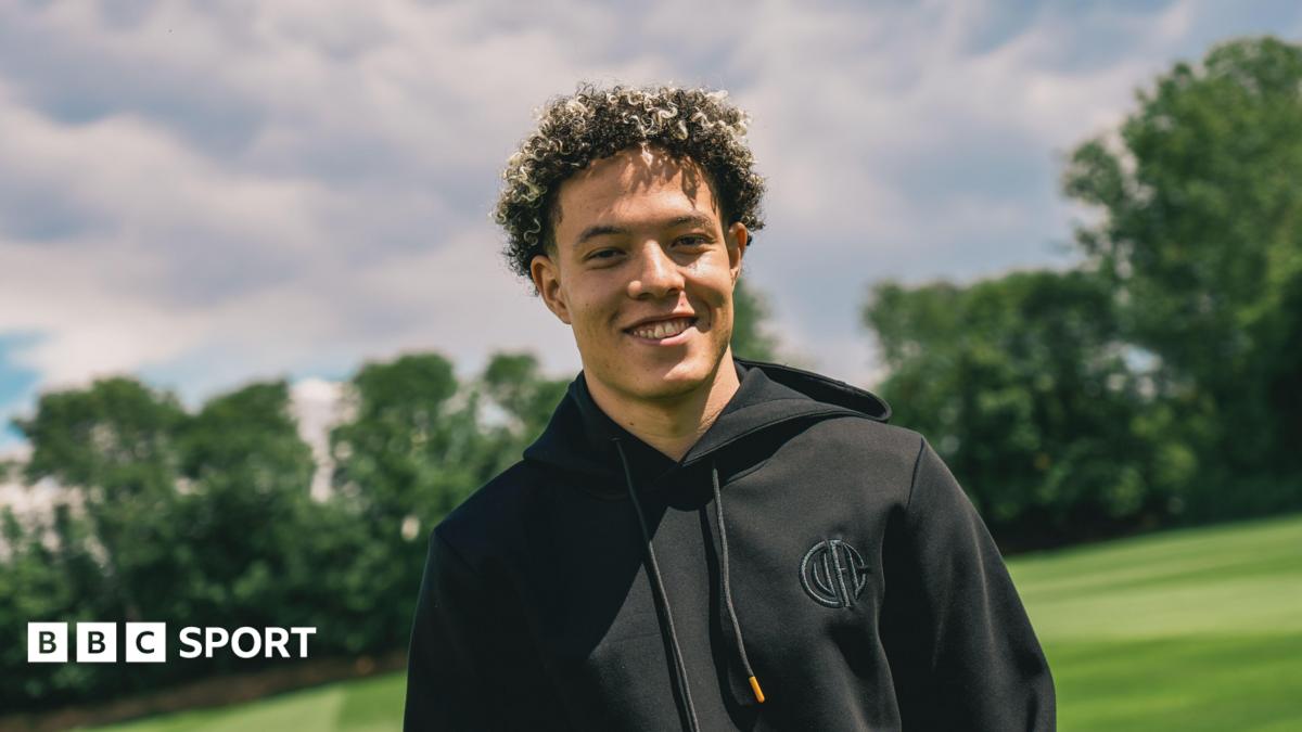 A photo of Isaac Heath smiling at the camera with trees in the background. He is wearing a black hoodie with the Cambridge United club badge on the chest
