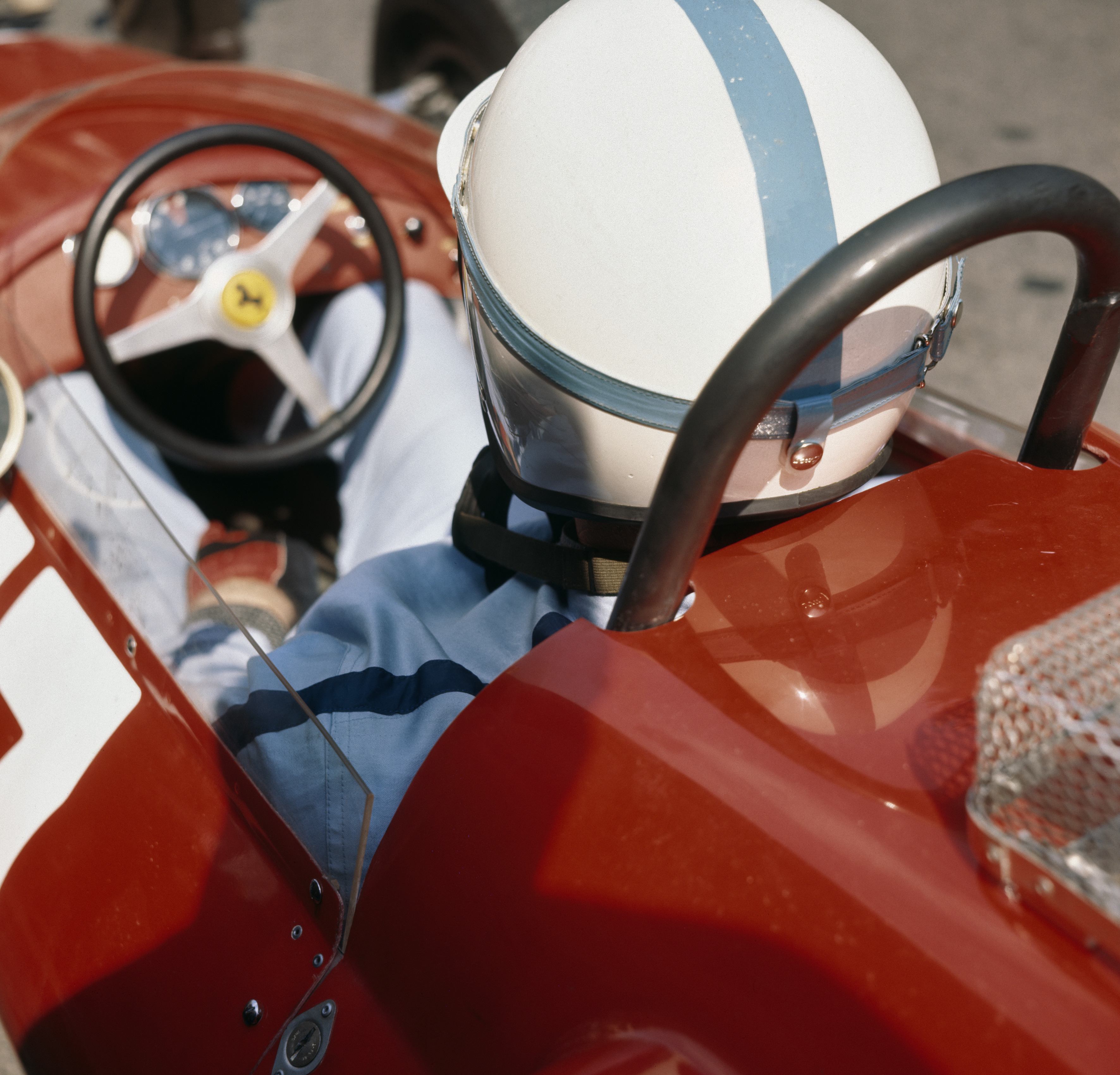 A racing driver in a white crash helmet with a blue stripe sits behind the wheel of a red Ferrari, photographed close-up from above.