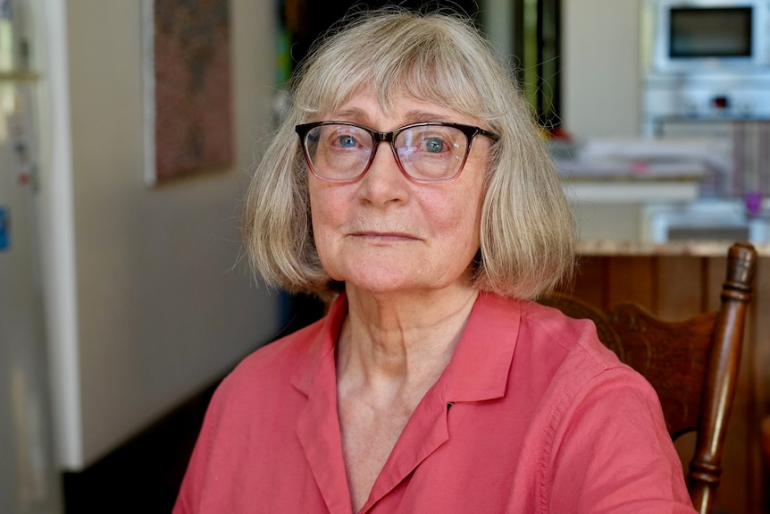 A woman with chin length light blonde hair, brown rimmed glasses in a pink shirt sits in a kitchen, staring at the camera.