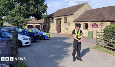 A uniformed police office stands in front of a bricked barn has a car park outside the front on the left. There is a fence on the right which opens into a court yard and there are signs on the door. There is a police car parked in the car park.