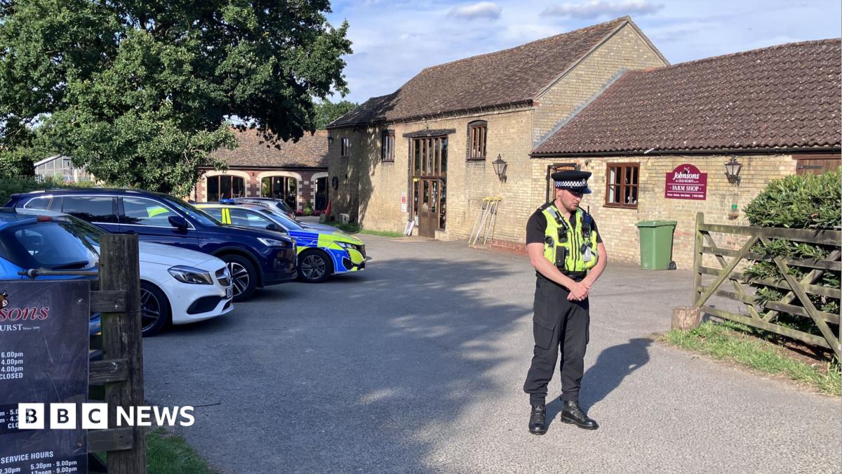 A uniformed police office stands in front of a bricked barn has a car park outside the front on the left. There is a fence on the right which opens into a court yard and there are signs on the door. There is a police car parked in the car park.