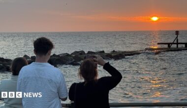 A view of a sunrise over a sea, watched by three people with phones held up filming it