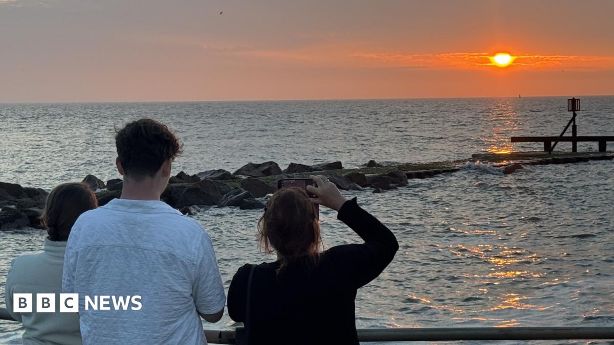 A view of a sunrise over a sea, watched by three people with phones held up filming it