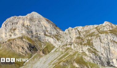 A large mountain with green vegetation at the bottom. The sky is bright blue behind it.