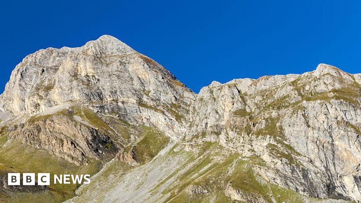 A large mountain with green vegetation at the bottom. The sky is bright blue behind it.