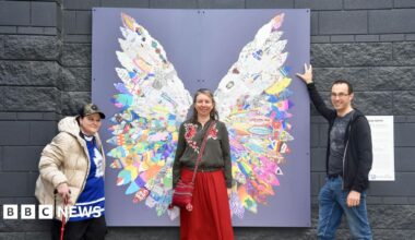 Three people stand around the artwork made of feathers that are placed in a way that makes them look like a pair of wings. One woman stands in the middle, as if they are her wings, and is smiling at the camera. A man and woman stand on either side of the piece, also smiling at the camera.