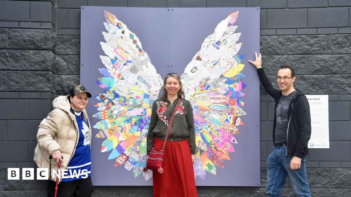 Three people stand around the artwork made of feathers that are placed in a way that makes them look like a pair of wings. One woman stands in the middle, as if they are her wings, and is smiling at the camera. A man and woman stand on either side of the piece, also smiling at the camera.