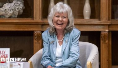 Dame Jilly Cooper laughs as she sits in a chair in front of a large, ornate, wooden shelving unit. She has grey hair to her shoulders and is wearing a blue satin suit jacket, a light blue V-neck top and a string of pearls around her neck. One of her books can be seen on a table in the foreground.