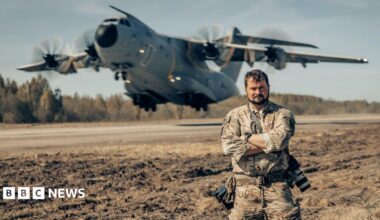 A man, Aaron Stone, stood with his armed crossed with two cameras hanging from his side. A large cargo plane is taking off in the background.