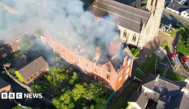 Drone footage from above of a red-brick building with flames inside it. The building is large and long with its roof caved in. It is surrounded by small red-brick buildings and a grey stone church beyond it. Firefighters in hi-viz protective clothing are spraying it with water.