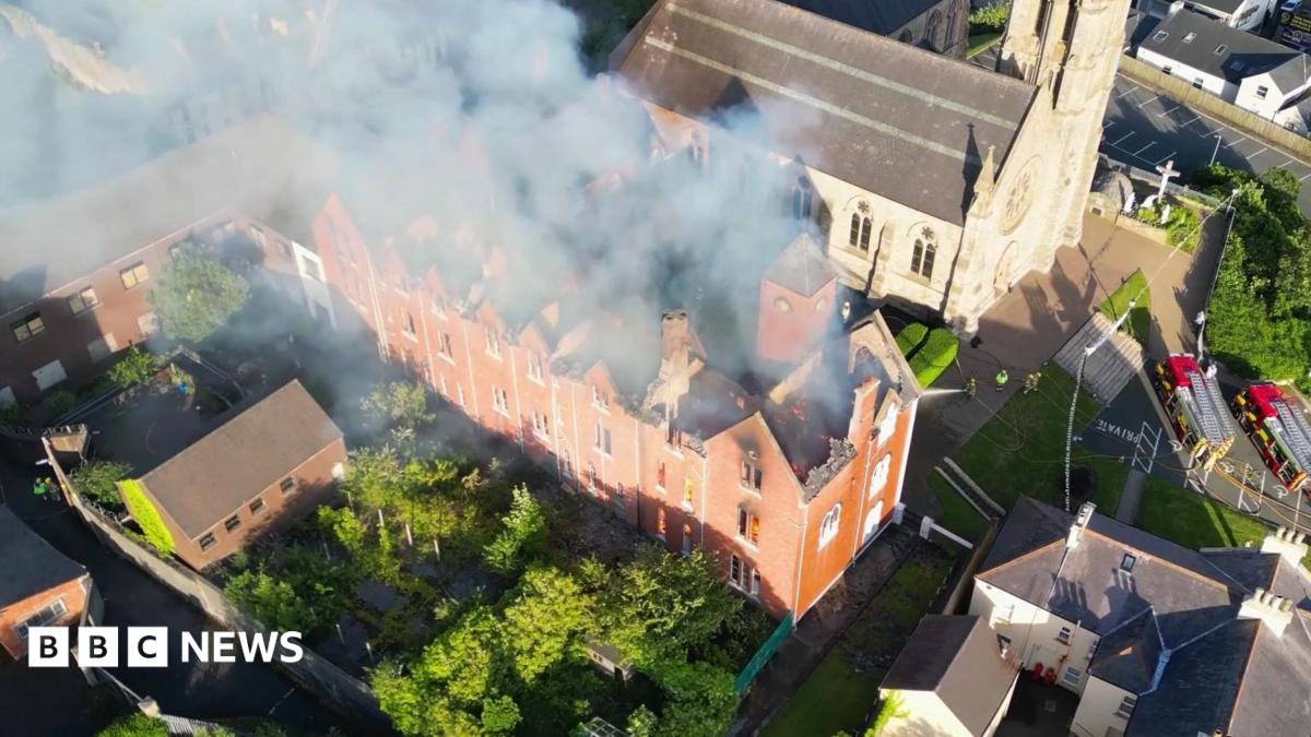 Drone footage from above of a red-brick building with flames inside it. The building is large and long with its roof caved in. It is surrounded by small red-brick buildings and a grey stone church beyond it. Firefighters in hi-viz protective clothing are spraying it with water.