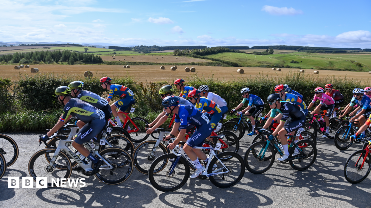 Several road cyclists dressed in various colours of lycra and helmets are all cycling down a country lane next to a field full of hay bales, with rolling fields behind it.