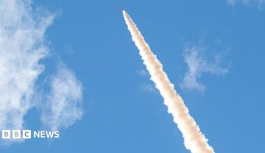 Smoke trails from the orange rocket as it heads up into a blue sky.