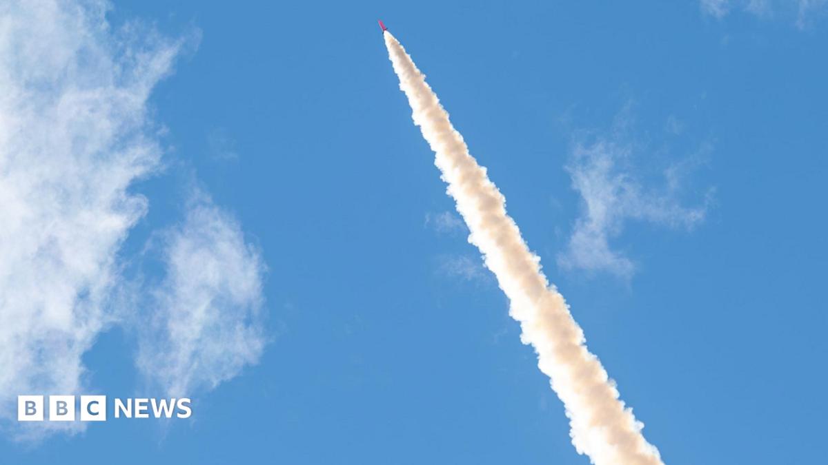 Smoke trails from the orange rocket as it heads up into a blue sky.