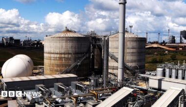 The sewage works which is made up of large metal structures which are rusting on the outside. The sky above is blue and full of fluffy white clouds.
