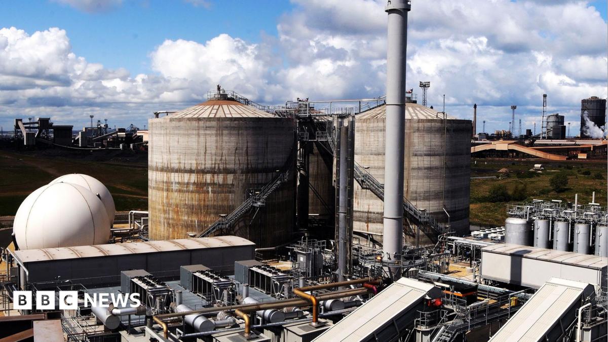 The sewage works which is made up of large metal structures which are rusting on the outside. The sky above is blue and full of fluffy white clouds.