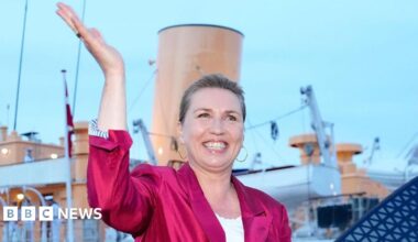 Danish Prime Minister Mette Frederiksen waves, smiling, in a bright pink blazer. She stands in front of the royal yacht Dannebrog.