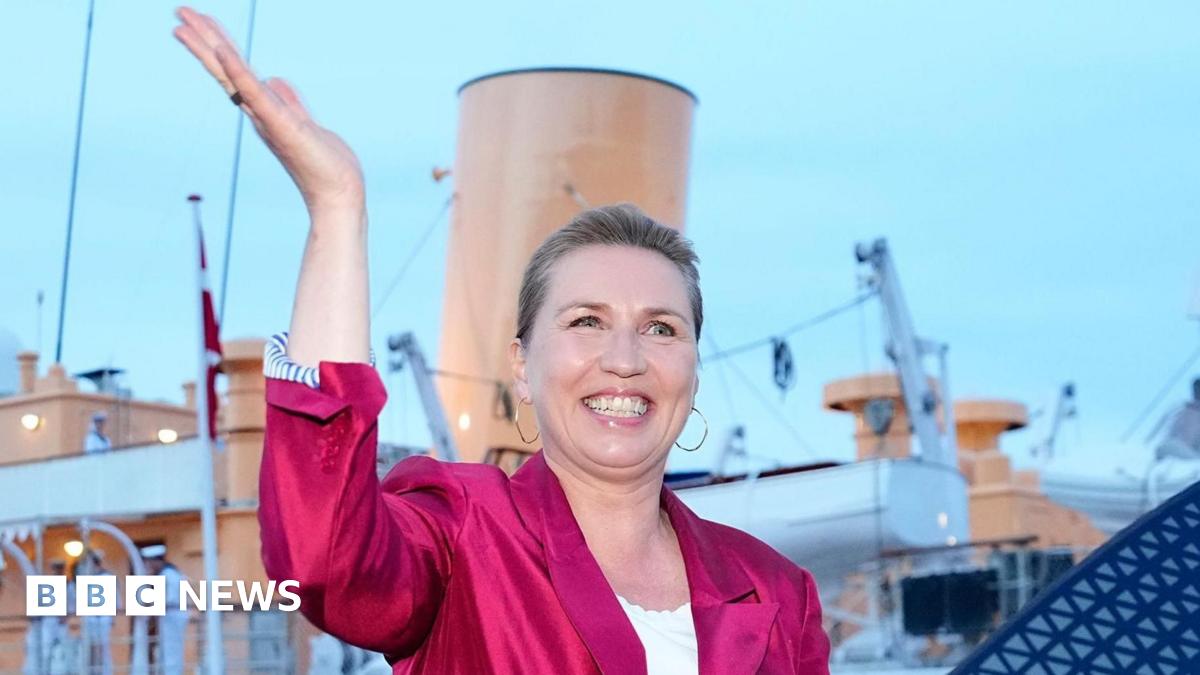 Danish Prime Minister Mette Frederiksen waves, smiling, in a bright pink blazer. She stands in front of the royal yacht Dannebrog.