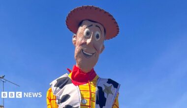 A little girl wearing a blue and white school dress stands in front of a wooden fence. A life-sized sculpture of Woody from the film Toy Story is sitting on top of the fence. He is wearing a brown cowboy hat, yellow and red chequered shirt, a jacket with a cow print on it and blue trousers.