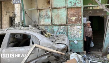 An elderly woman stands in the entrance to a damaged apartment block. To her left is a badly damaged car with no windows. Debris lies all around.