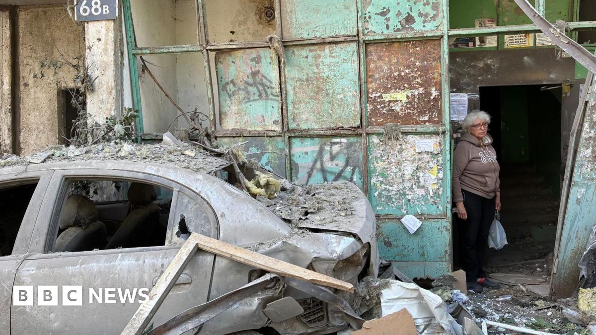 An elderly woman stands in the entrance to a damaged apartment block. To her left is a badly damaged car with no windows. Debris lies all around.