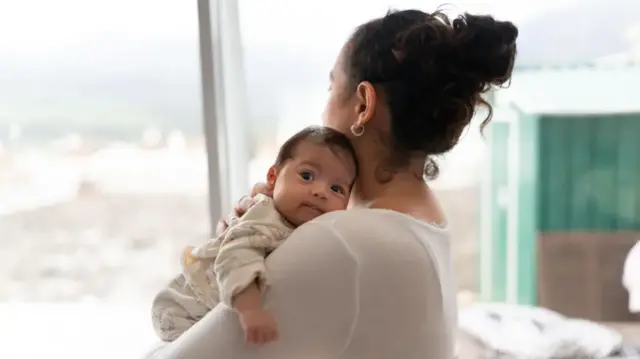 A woman in a white shirt looks out of a window whilst holding her baby